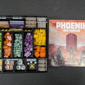 Puede incluir: Un organizador de caja de juego con múltiples compartimentos para guardar las piezas y las cartas del juego. El organizador está lleno de piezas de plástico de varios colores y cartas. La tapa de la caja está abierta y muestra el reglamento del juego con el título "PHOENIX NEW HORIZON".