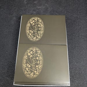 May include: Two rectangular boxes with a dark gray exterior and white edges. Each box features an oval design with the word "Indonesia" in gold script surrounded by floral and leaf patterns. The boxes are stacked on top of each other.