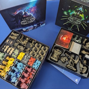 Puede incluir: Una caja de juego negra y verde con el título "Lords of Ragnarok Stretch Goals" impreso en la tapa. La caja está abierta y contiene un inserto de espuma con compartimentos para piezas de juego, cartas y fichas. Las piezas son principalmente marrones y grises, con algunos acentos rojos y amarillos.
