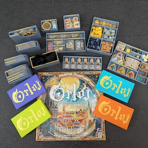 May include: A board game titled "Orloj: The Prague Astronomical Clock" with various game pieces and organizers. The game box features a detailed illustration of a clock tower. Several rectangular game pieces are in blue, orange, green, and purple.