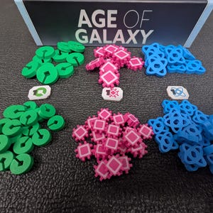 Puede incluir: Un juego de mesa titulado "AGE OF GALAXY" con varias piezas. Las piezas son verdes, rosas y azules, con formas únicas. También hay pequeñas fichas blancas con símbolos. Las piezas del juego están dispuestas sobre una superficie oscura.