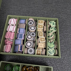 May include: A green organizer box filled with game pieces. The pieces are various shapes and colors, including pink, blue, gray, brown, and green. The box is divided into compartments for easy organization.