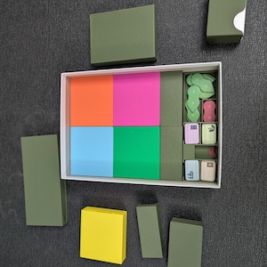May include: A white box with a variety of colorful blocks and small, decorative items. The blocks are orange, pink, blue, and green. The small items are green, pink, and beige. Several olive green boxes are arranged around the white box.