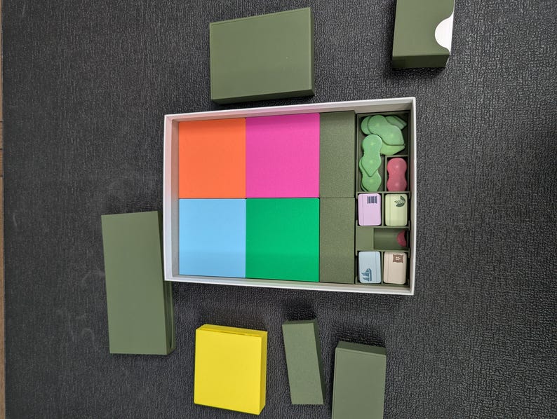 May include: A white box with a variety of colorful blocks and small, decorative items. The blocks are orange, pink, blue, and green. The small items are green, pink, and beige. Several olive green boxes are arranged around the white box.