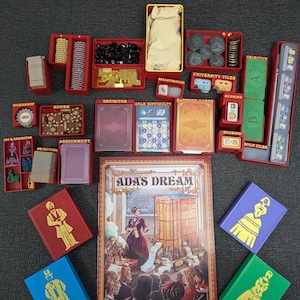 May include: A board game set with various components, including red boxes, cards, tokens, and a rulebook titled "ADA'S DREAM." The rulebook features an illustration of a woman and a complex machine. Other components include tiles and tokens.