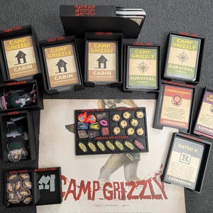 May include: A Camp Grizzly board game set featuring multiple black plastic card organizers, decks labeled Cabin, Survival, Cheerleaders, Behind the Bear, and Rated R, a central token tray with various game pieces, and a game board with the title text.