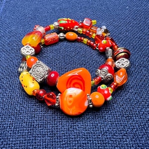 May include: A handmade bracelet featuring a variety of colorful beads in shades of orange, red, yellow, and silver. The beads are of different shapes and sizes, including round, oval, and irregular. The bracelet is on a blue background.