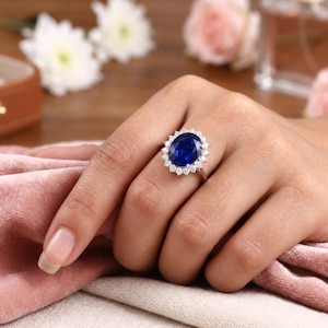 Könnte beinhalten: Ein silberner Ring mit einem großen, oval geschliffenen, tiefblauen Edelstein in der Mitte, umgeben von einem Kranz aus kleinen, runden, klaren weißen Steinen. Die Ringschiene ist dünn und metallisch und zeigt ein klassisches Design mit einem blauen Fokus.