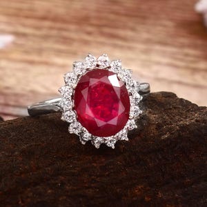 May include: A silver ring featuring a large, oval-cut ruby gemstone surrounded by a halo of smaller, clear gemstones. The ruby is a deep red color, and the ring is set against a dark, textured background.