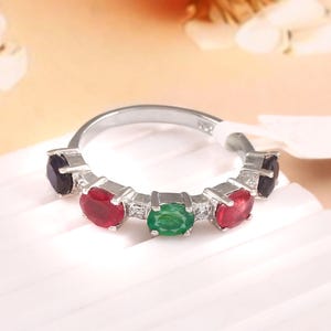 Può includere: Un anello d'argento con una fila di pietre preziose. L'anello presenta una pietra verde centrale, due pietre rosse su ciascun lato e una pietra viola scuro a ciascuna estremità. L'anello è su sfondo bianco.