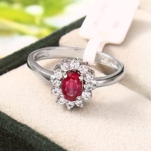 May include: A silver ring with an oval ruby centre stone, encircled by smaller clear gemstones. The ring is set in a silver band and presented in a dark green jewellery box. The ring is displayed with a white tag.