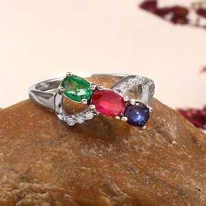 May include: A silver ring with three oval gemstones in green, red, and blue. The ring has a distinctive design with small, clear accent stones. The ring is set on a brown stone surface.