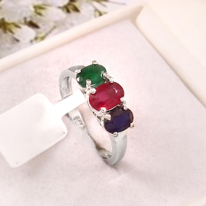 Puede incluir: Anillo de plata con tres piedras preciosas ovales: verde, rojo y azul. Las piedras están dispuestas en fila, creando un diseño colorido y llamativo. El anillo se presenta en una caja de joyería blanca.