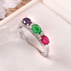 Puede incluir: Un anillo de plata con tres piedras preciosas ovales en fila: un zafiro azul oscuro, una esmeralda verde y un rubí rojo. Pequeñas piedras transparentes se colocan entre las piedras preciosas. El anillo se muestra en una caja blanca.