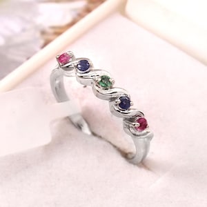 Könnte beinhalten: Ein silberner Ring mit wellenförmigem Design, besetzt mit abwechselnden Edelsteinen. Der Ring hat kleine, runde Edelsteine in Rot, Blau und Grün. Der Ring wird in einer weißen Schmuckschatulle präsentiert.