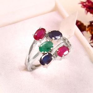 May include: A silver ring featuring six oval gemstones in shades of red, green, and blue. The gemstones are set in a swirling design, creating a unique and eye-catching piece of jewelry. The ring is displayed in a white box.