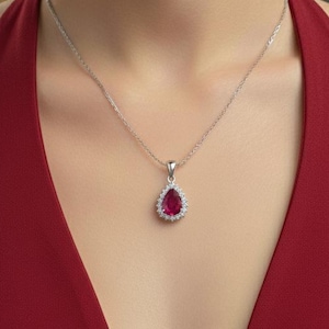 Pode incluir: Um colar de prata com um pingente em forma de gota. O pingente apresenta uma grande pedra preciosa vermelha rodeada por pedras claras menores. O colar é usado com um vestido vermelho.