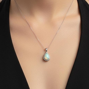 May include: A silver necklace with a teardrop-shaped pendant. The pendant features a central opal stone surrounded by a halo of small, clear gemstones. The necklace is worn against a black dress.