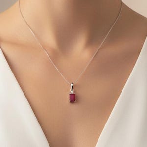 May include: A silver necklace featuring a rectangular, deep red gemstone pendant suspended from a delicate chain. The pendant is set in a simple silver frame, resting against a plain white sleeveless garment with a V-neckline.