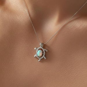 Op de afbeelding: Een zilveren ketting met een schildpadhanger. De hanger heeft een ovale, opalescente centrale steen, omringd door kleine, heldere edelstenen. De ketting wordt getoond op de hals en borst van een persoon.
