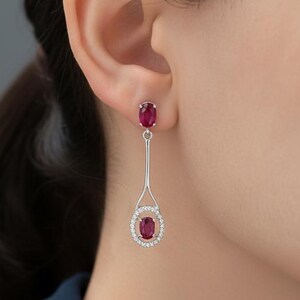 May include: Elegant silver earrings with oval ruby gemstones. The earrings feature a small ruby stud at the ear and a dangling design with a larger ruby surrounded by a halo of small, sparkling stones.