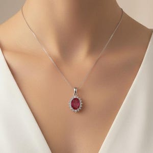 May include: A silver pendant necklace featuring an oval, deep red gemstone surrounded by small, clear stones. The pendant hangs from a delicate silver chain. The necklace is displayed against a white dress.