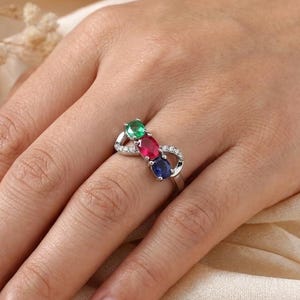 Könnte beinhalten: Ein silberfarbener Ring mit einem unendlich geformten Band, das mit drei oval geschliffenen Edelsteinen in Grün, Rot und Blau besetzt ist, akzentuiert durch kleine klare Steine an den Seiten des Bandes, was ein poliertes metallisches Aussehen ergibt.