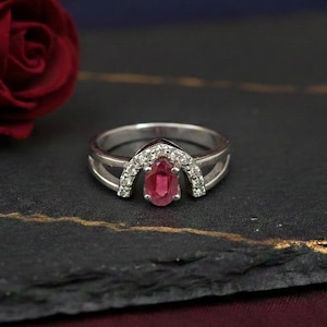 May include: A silver ring featuring a pear-shaped, red gemstone, encircled by smaller white stones. The ring is set against a dark surface with a red rose in the background. The ring has a split shank design.