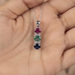 May include: A silver-toned necklace featuring a vertical pendant with three round-cut gemstones in red, green, and dark blue, accented by small clear stones set in a metal frame, suspended from a delicate silver-toned chain.