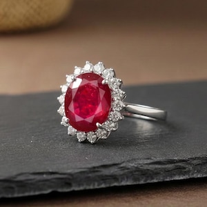 Könnte beinhalten: Ein silberner Ring mit einem großen, oval geschliffenen Rubin, umgeben von kleineren, klaren Diamanten. Der Rubin hat eine tiefe rote Farbe, und die Diamanten funkeln. Der Ring ist ein klassisches Design, geeignet für besondere Anlässe.