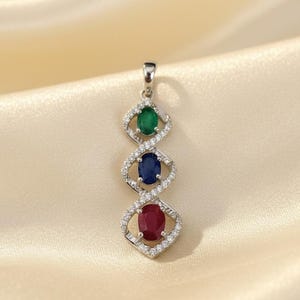 Puede incluir: Colgante de plata con tres piedras preciosas ovales: una esmeralda verde, un zafiro azul y un rubí rojo. Cada gema está engastada en un marco en forma de rombo con diamantes. El colgante cuelga de un pequeño lazo.