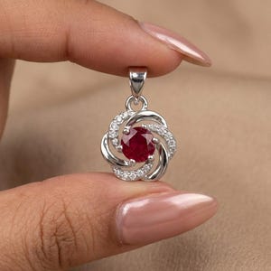 Puede incluir: Un colgante de plata con una piedra preciosa redonda de color rojo intenso en el centro, rodeada por un marco circular en espiral incrustado con pequeñas piedras blancas transparentes. El colgante incluye una reasa de plata en la parte superior.