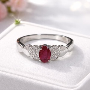 May include: A silver-toned ring featuring a central oval-cut deep red ruby gemstone. The ruby is flanked on each side by a small triangular cluster of three clear, round-cut accent stones set into the polished metal band of the ring.