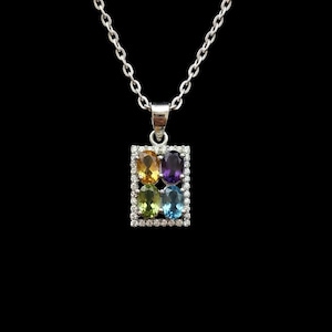 Puede incluir: Un collar de plata con un colgante rectangular que presenta cuatro piedras preciosas de corte ovalado en amarillo, morado, verde y azul. El colgante está bordeado por pequeñas piedras blancas y cuelga de una fina cadena de plata sobre fondo negro.