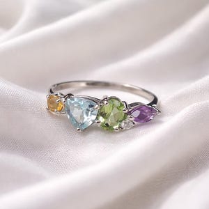 Könnte beinhalten: Ein silberner Ring mit einer Reihe farbiger Edelsteine. Der Ring zeigt einen gelben, hellblauen, grünen und lila Edelstein. Die Edelsteine sind in einem silbernen Band gefasst und bilden ein lebendiges und auffälliges Schmuckstück.