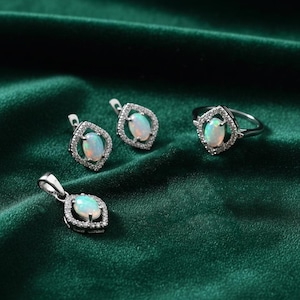 May include: A jewellery set comprising an opal ring, pendant, and earrings. Each piece has a silver setting with an oval opal stone at the centre, encircled by small, clear accent stones. The jewellery is displayed on a dark green velvet surface.