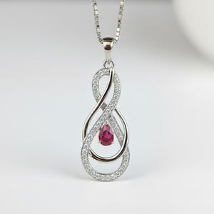 Può includere: Una collana con pendente in argento con un design a simbolo dell'infinito. Il pendente è adornato con piccole gemme trasparenti e una gemma rosso scuro a forma di goccia. La collana è appesa a una delicata catena d'argento.