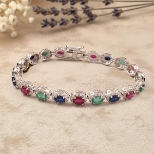 Könnte beinhalten: Ein silbernes Armband mit oval geschliffenen Edelsteinen in Rot, Grün und Blau. Die Edelsteine sind in einer dekorativen Silberfassung mit kleinen, klaren Steinen gefasst. Das Armband hat einen Verschluss.
