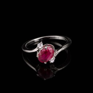 May include: A silver ring featuring a large, oval-cut ruby gemstone. The ruby is a deep red color and is accented by smaller, clear gemstones on either side. The ring is set against a black background, creating a striking contrast.
