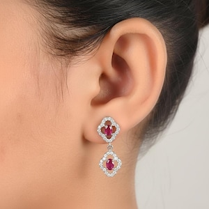Pode incluir: Brincos pendurados com um design em forma de trevo e uma pedra preciosa de rubi central. Os brincos são rodeados por um halo de pequenas pedras brancas brilhantes, criando um visual luxuoso. Eles são presos à orelha com um pino.