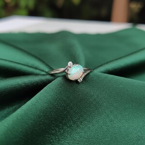 May include: A silver ring with an oval opal gemstone. The opal has a milky white and light blue color. The ring has small accent stones on either side of the opal. The ring is set against a dark green fabric.