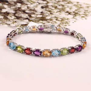 Può includere: Un bracciale in argento con una serie di pietre preziose ovali di vari colori. Le pietre sono disposte in fila, creando un gioiello vivace. Il bracciale ha una chiusura sicura.