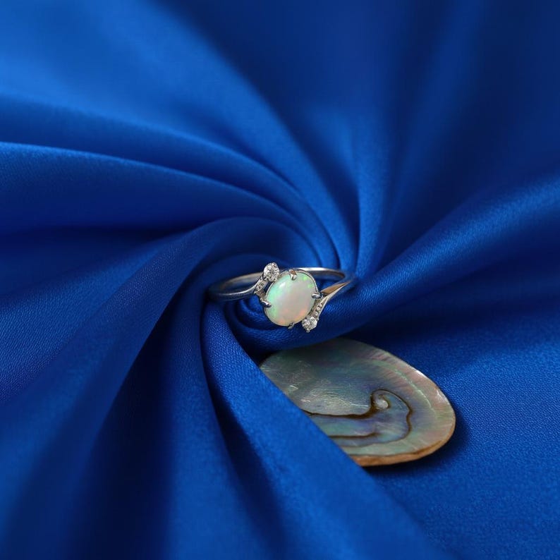 May include: A silver ring with an oval opal gemstone and small accent stones. The ring is set against a vibrant blue fabric, with a pearlescent shell in the background. The ring's design is delicate and elegant.