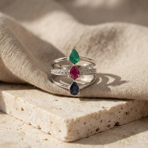 May include: A silver ring featuring three stacked bands, each set with a pear-shaped gemstone. The top stone is green, the middle stone is red, and the bottom stone is dark blue. The middle band is accented with small, clear, round stones.