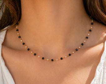 May include: A delicate gold chain necklace featuring an alternating pattern of small, round black beads and tiny white pearls. The necklace rests against the neck, showcasing a simple link design that connects each bead and pearl individually.