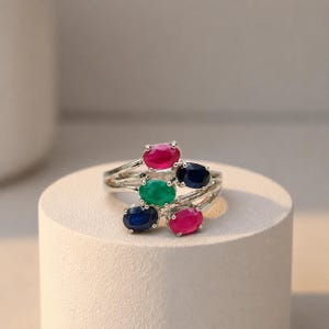 May include: A silver-toned ring featuring a multi-strand band set with five oval-cut gemstones. The arrangement includes two vibrant pink stones, two deep blue stones, and one central green stone, all secured in prong settings.