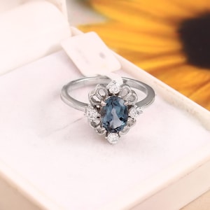 May include: A silver ring with an oval, blue gemstone center, surrounded by small, clear accent stones. The ring's setting has a decorative, openwork design. The ring is displayed in a white jewelry box, with a yellow and orange background.