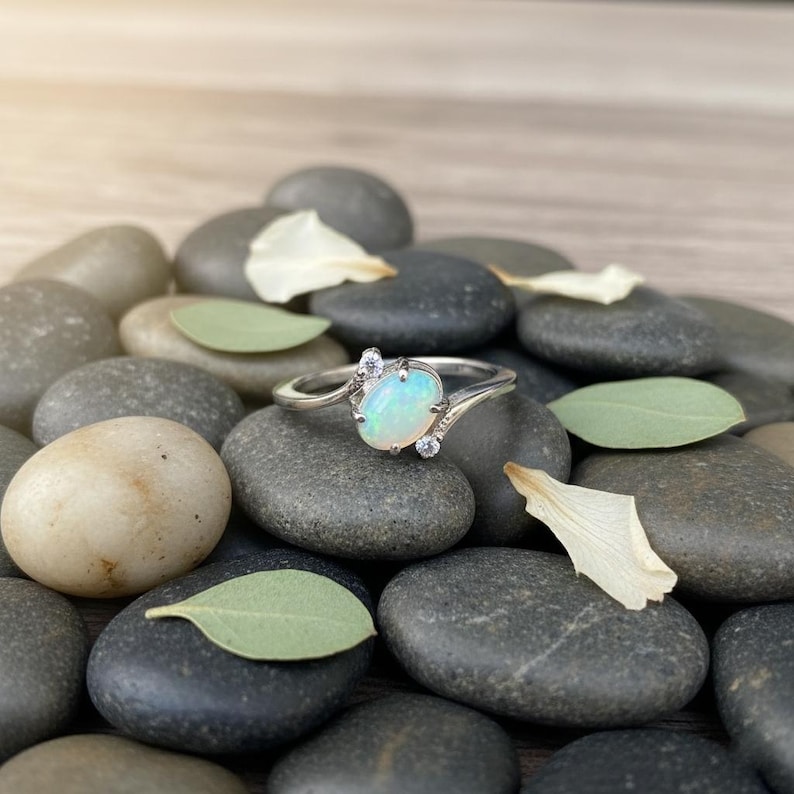 May include: A silver ring with an oval opal gemstone and small accent stones. The ring is set on a bed of smooth, gray stones with scattered leaves and petals. The opal has a milky, iridescent appearance.
