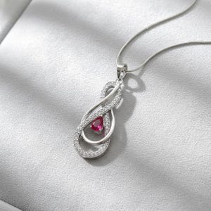 Peut inclure: Un collier pendentif argenté avec un symbole infini. Le pendentif présente une pierre précieuse rouge en forme de goutte d'eau au centre, entourée de petites pierres claires. La chaîne du collier est fine et argentée.