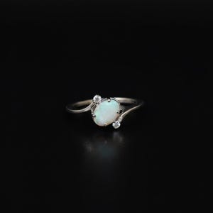May include: A silver ring featuring an oval opal gemstone and two smaller accent stones. The opal has a milky white appearance. The ring's band curves gracefully around the main stone, with the accent stones set on either side.